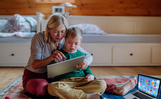 grandmother playing with a child with diverse abilities on a tablet with laptop on the floor.